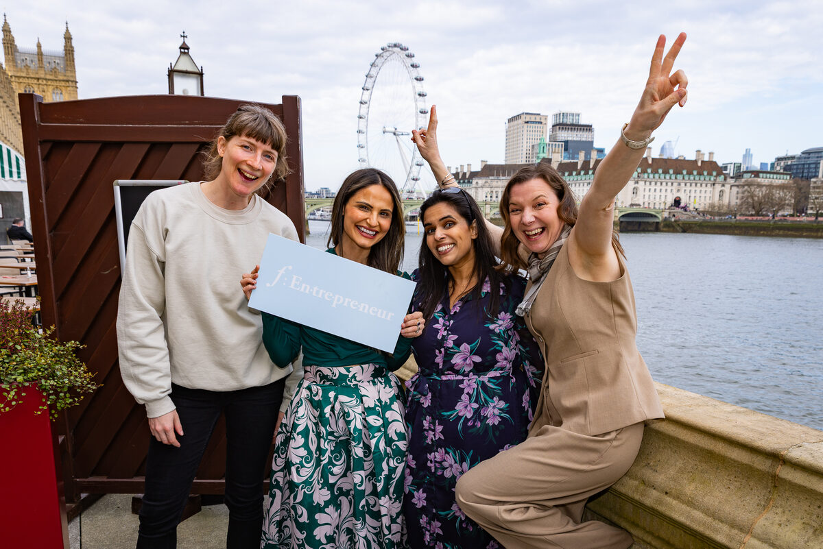 Four Female Founders List members holding an f:Entrepreneur sign beside the River Thames, with the London Eye behind them